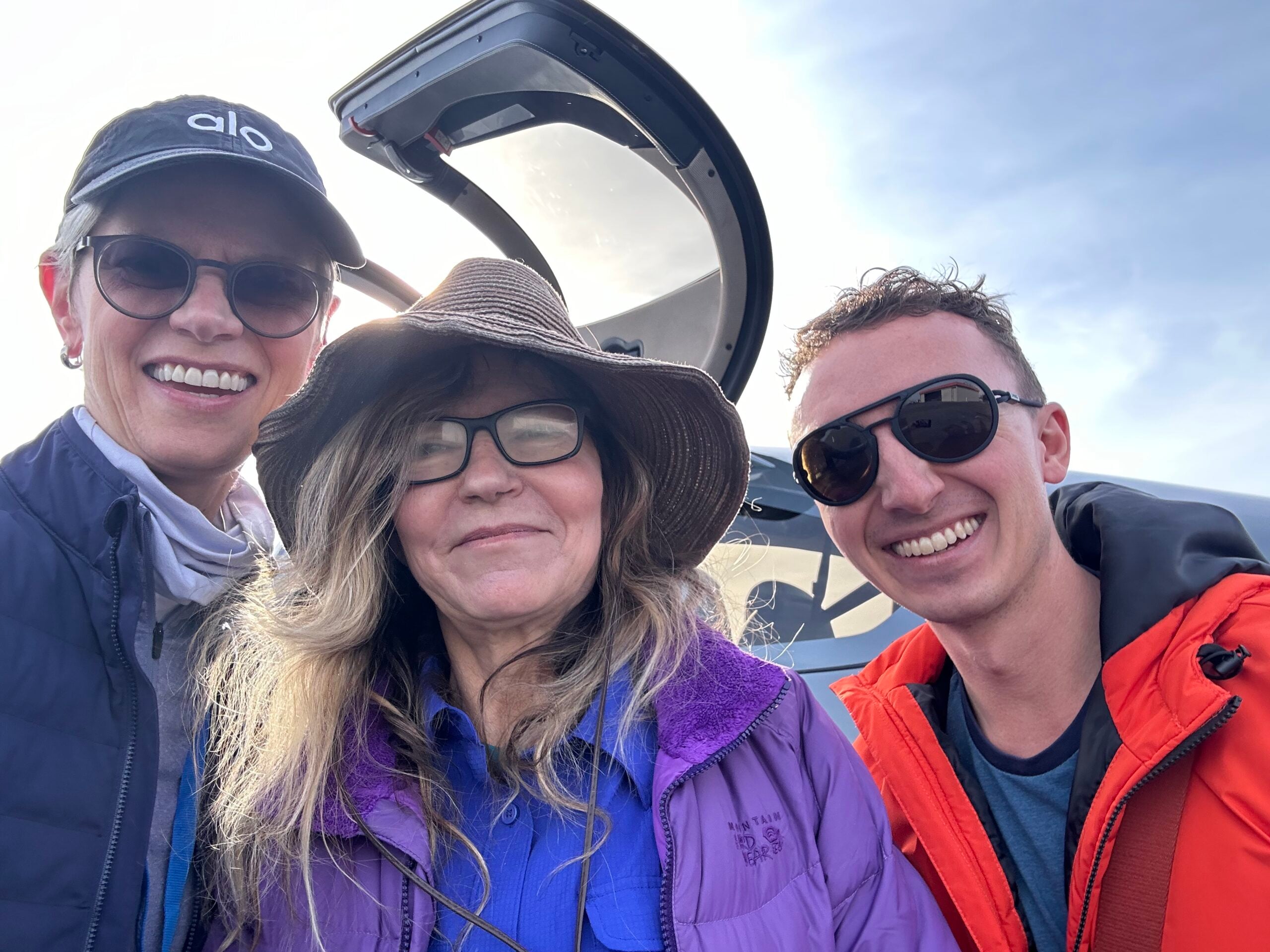 Three people smiling for a selfie beside a small plane with its door open on a sunny day.