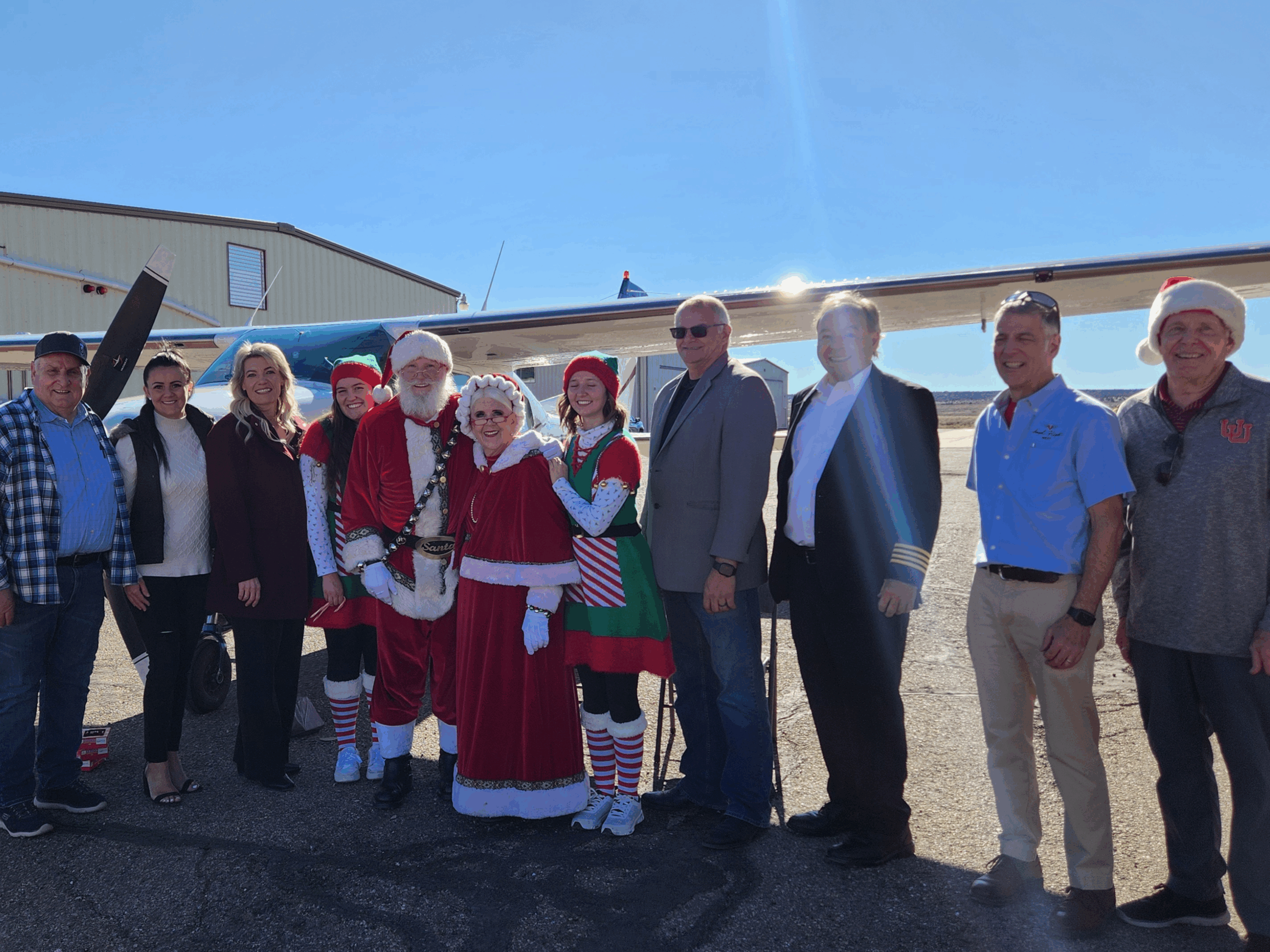 A group including Santa, Mrs. Claus and two elves stands in a row on an airport ramp in front of a small plane.