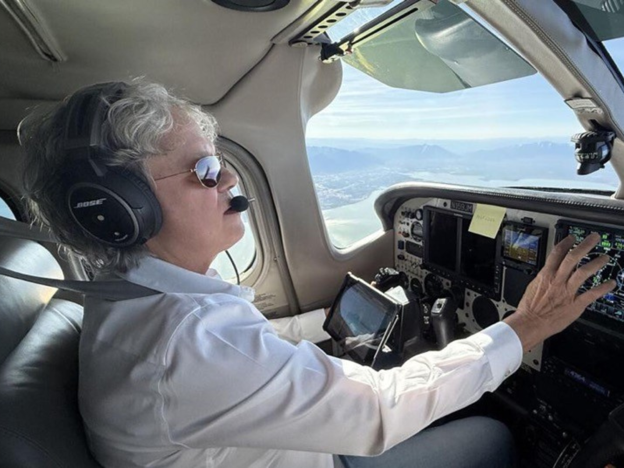 Pilot wearing Bose headset and sunglasses, seated in a small aircraft cockpit and adjusting the controls while looking at the instruments.