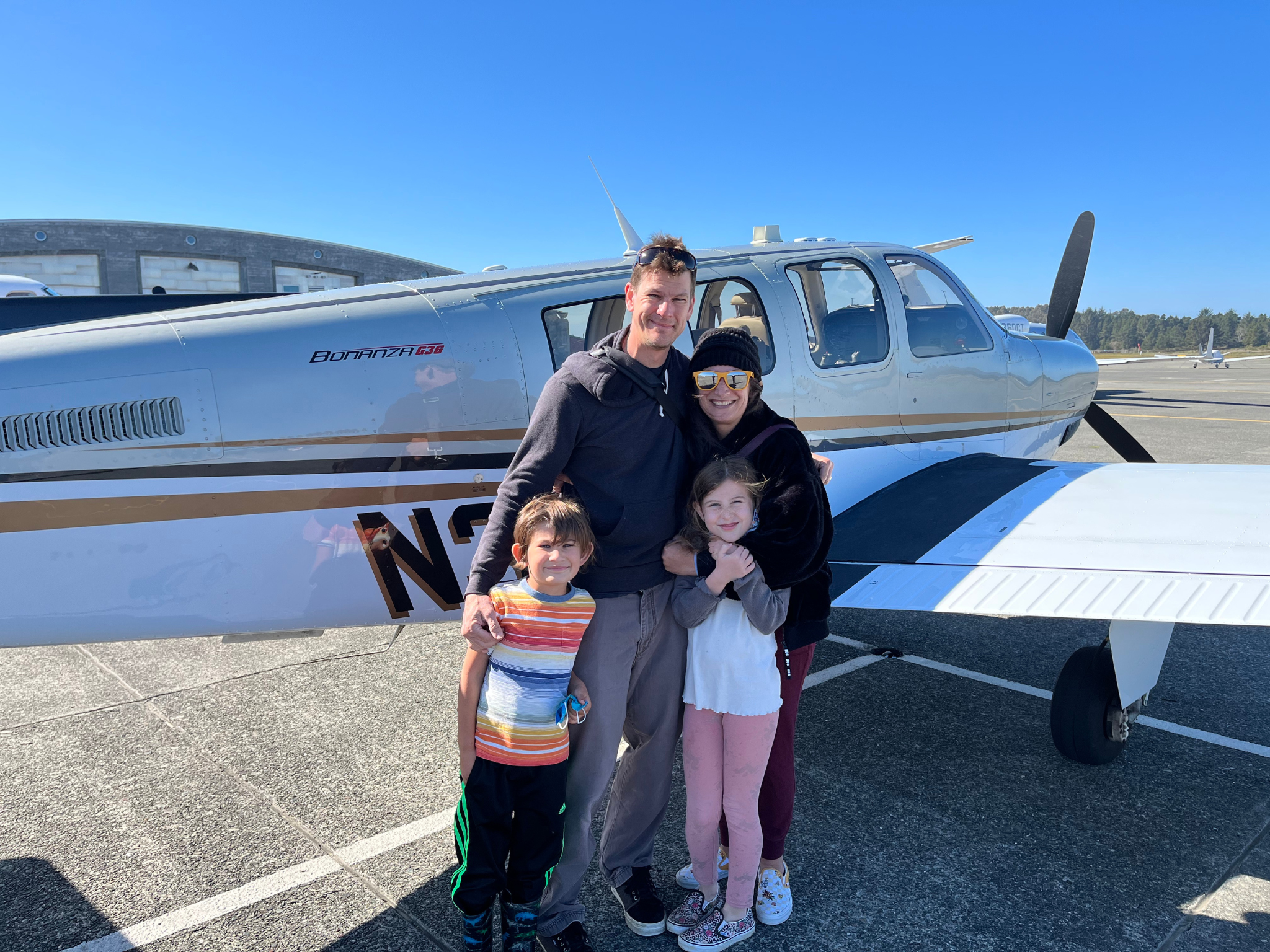 Family posing in front of a silver single-engine airplane on a sunny tarmac, blue sky overhead.