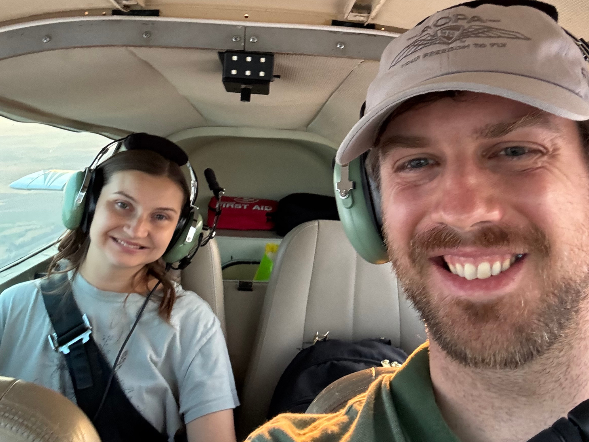 Smiling man in the foreground and a woman in the background wearing headsets in a small airplane cockpit.