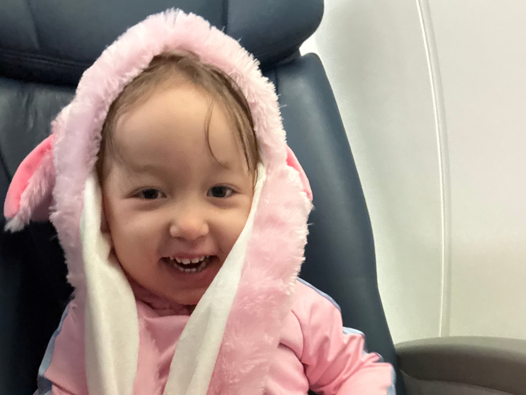 Baby in a pink fluffy hooded onesie smiling at the camera and showing teeth.