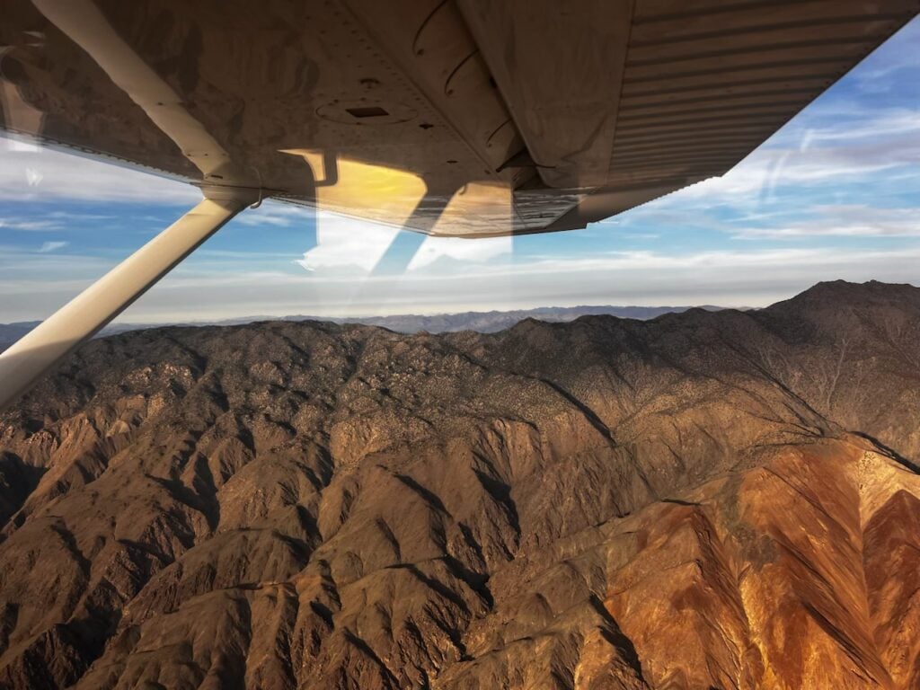 A small airplane wing and strut over rugged brown mountains seen from above, with a blue sky and canyon shadows below.