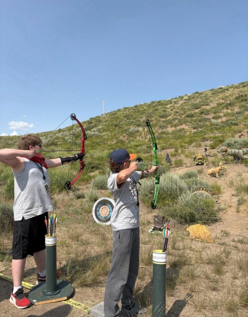 Two kids practice archery outdoors, drawing bows toward targets with dinosaur figures in the desert background.&nbsp;