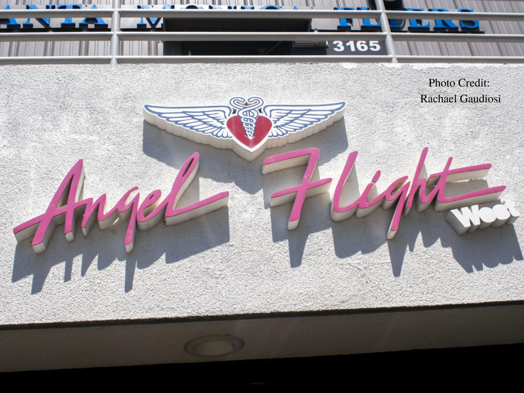 Exterior wall with a pink 'Angel Flight' sign and winged-heart logo; photo credit on the right side.