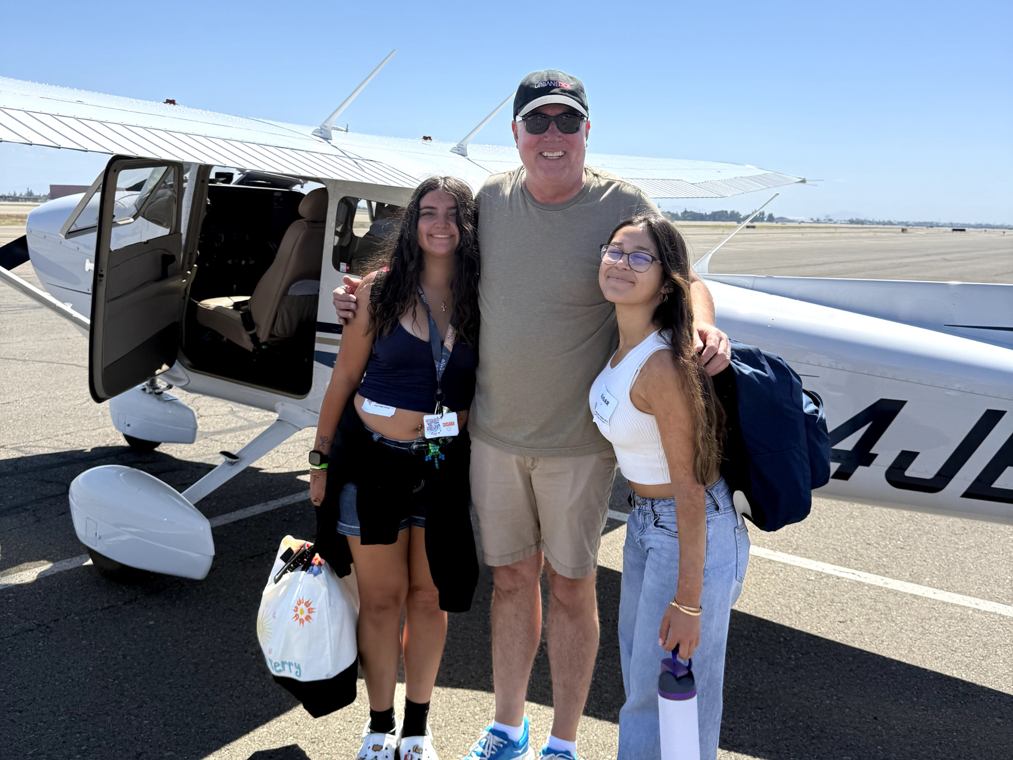 Three people posing beside a small white airplane with its door open on a sunny tarmac.