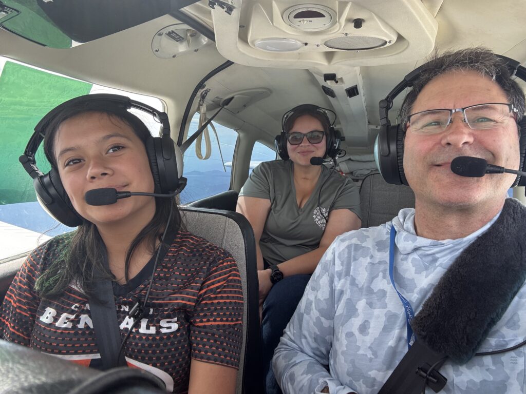 Three people wearing aviation headsets sit in a small airplane cockpit: a man on the right, a girl on the left, and a woman in the middle smiling softly.&nbsp;