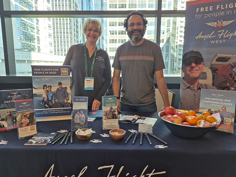 Man and woman standing behind an Angel Flight West table filled with marketing materials at Cancer Con (Convention).