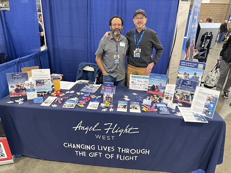 Two men, Tevor and Ricardo, standing together, smiling, behind a table at a convention with marketing supplies on top of the table.