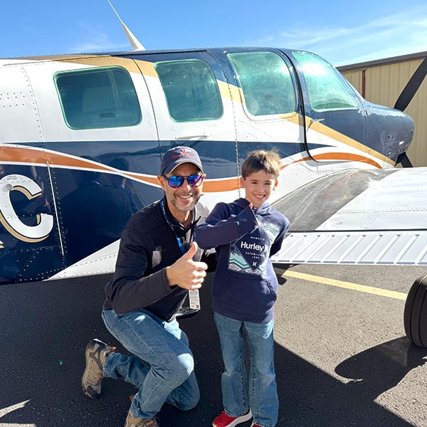 Man with a thumbs up and young boy smiling, standing in front of a small airplane looking at the camera.