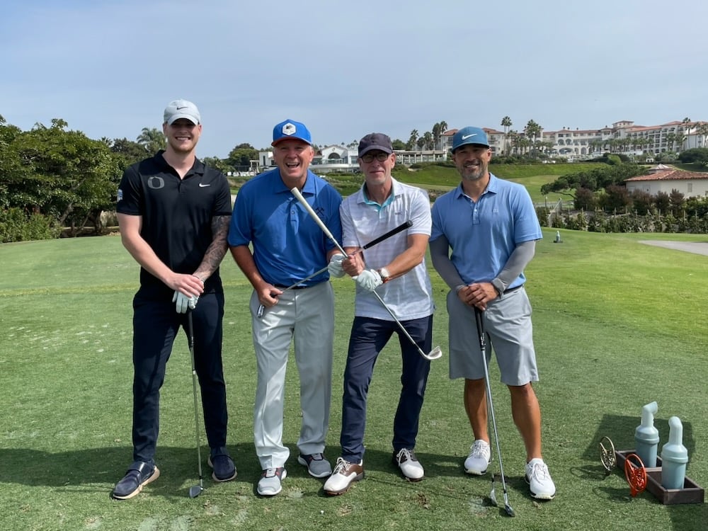 4 men standing together smiling for a picture at a golf course holding their clubs in an "x" formation