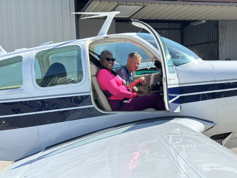 mission_photo_2023-10-14_James_Koritz_10253 - Angel Flight West Smiling couple seated in a small airplane with open door, parked near a hangar (woman in pink top, man beside her).