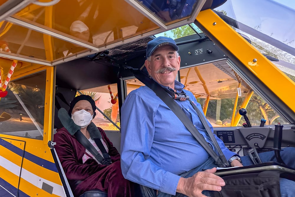 Pilot sitting in the front of the airplane with a patient sitting behind him wearing a mask, both looking at the camera.