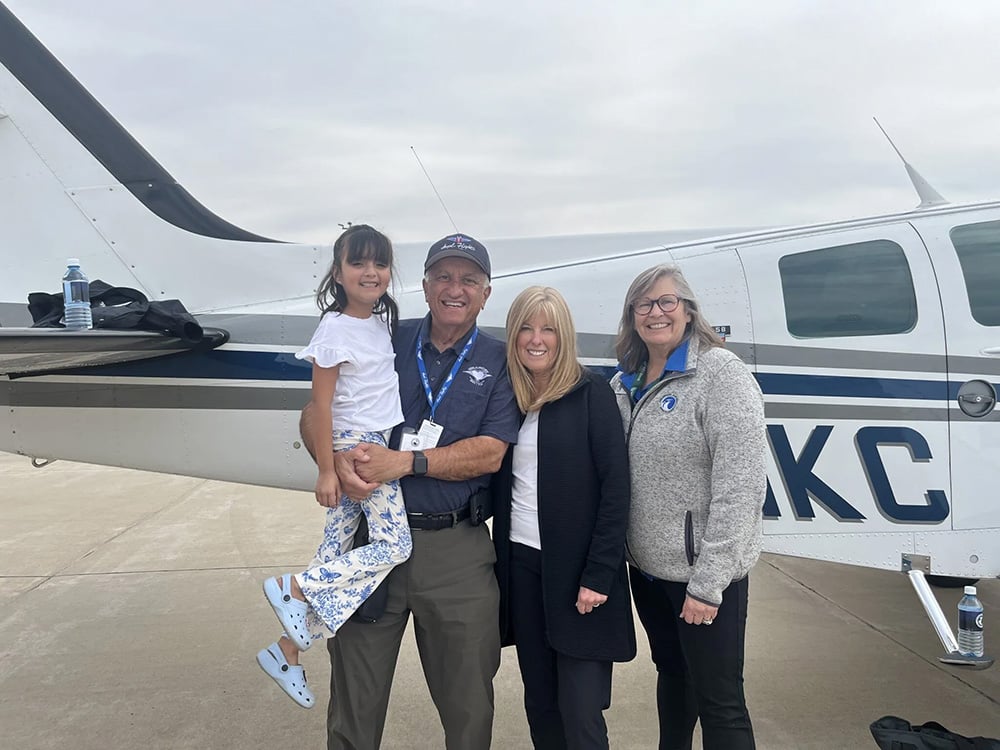 Group of people with a young girl in the older mans arms standing next to a small airplane smiling for the picture.