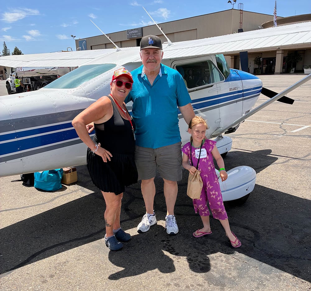 A small child wearing a pink outfit with her mom and dad standing in front of a small airplane smiling at the camera.