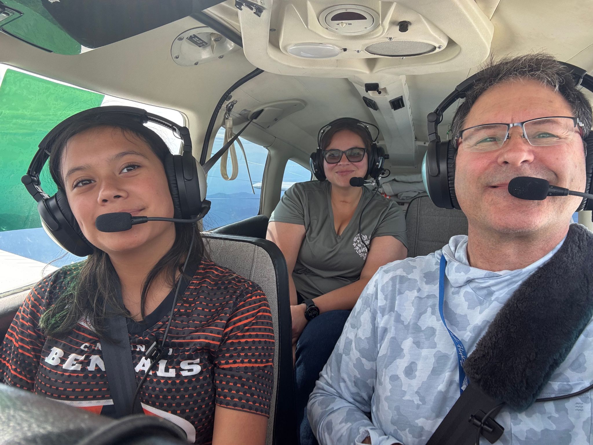 A pilot and two passengers in headsets smile from inside the cabin of a small plane.