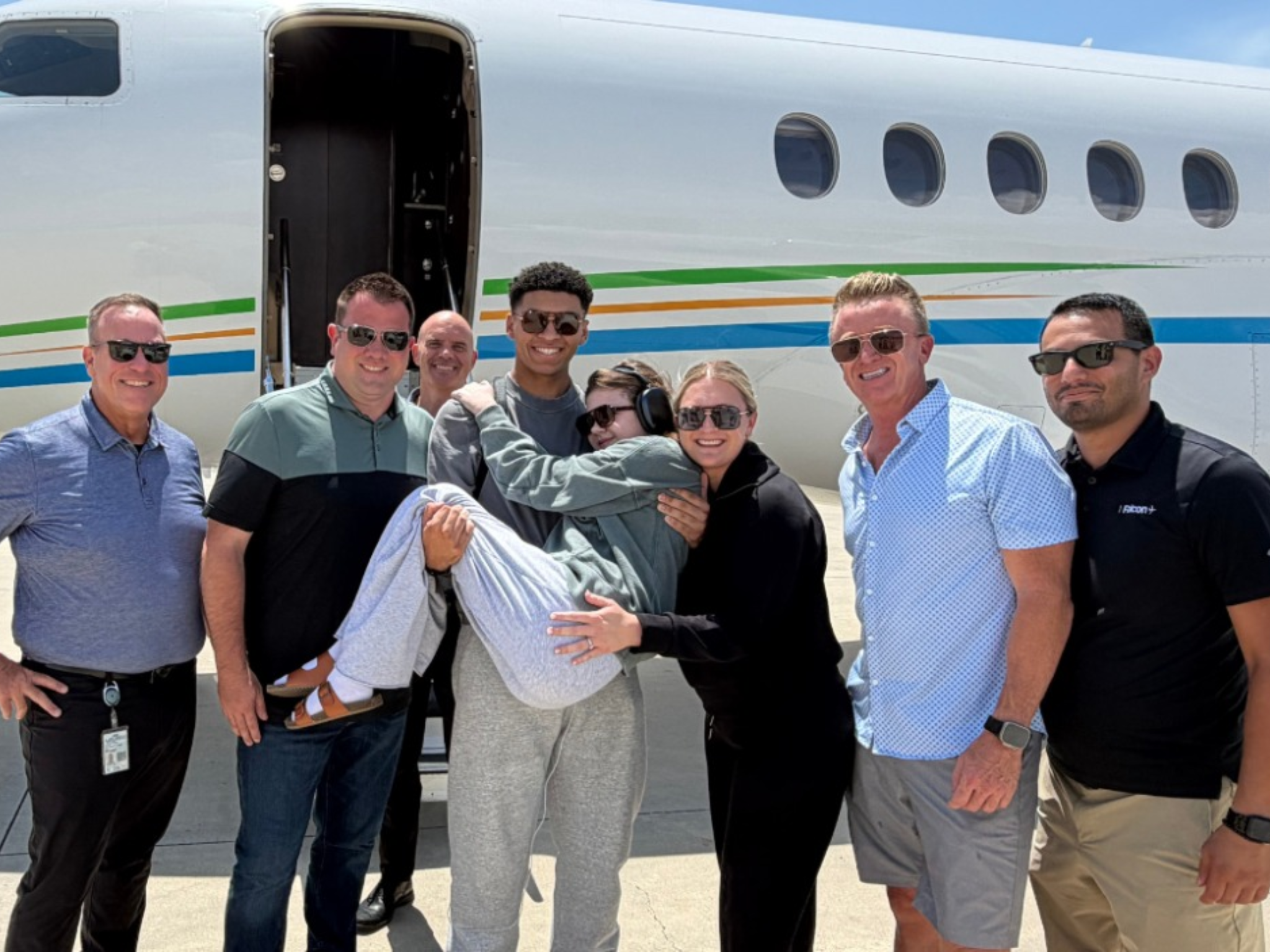 A group stands smiling in front of a private jet while one man carries a young passenger in his arms.